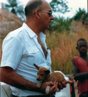 Man cradling a red basenji puppy in his arms, a boy standing behind him in the grass