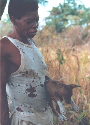 Woman in a white tank top carrying a red basenji puppy through tall grass