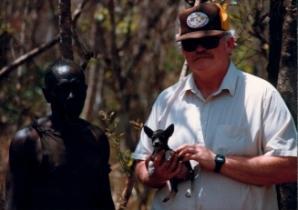 Man in a cap holding a small basenji puppy, standing beside a villager in the bush